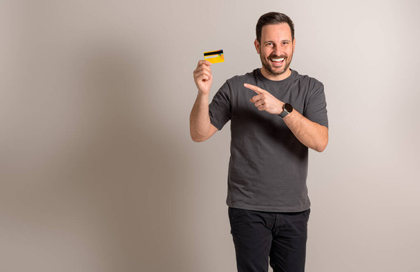 Portrait of shopaholic young man showing and pointing at credit card against background. Handsome male customer laughing and recommending online shopping and electronic banking - Fotó, kép