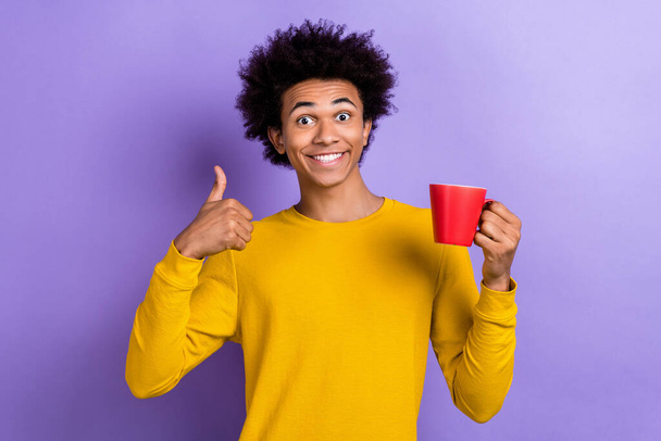 Photo of optimistic guy dressed yellow long sleeve hold cup of tea showing thumb up good taste isolated on purple color background. - 写真・画像