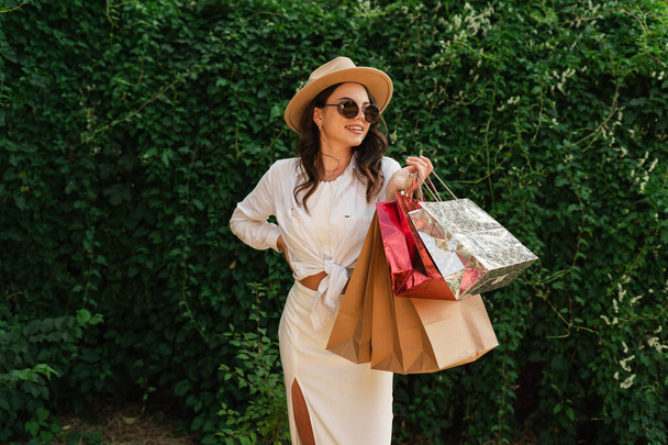 Joyeux jeune femme porter des vêtements blancs et chapeau de paille tenir des sacs de paquet après les achats isolés sur fond de verdure. Concept de repos estival. Espace de copie - Photo, image