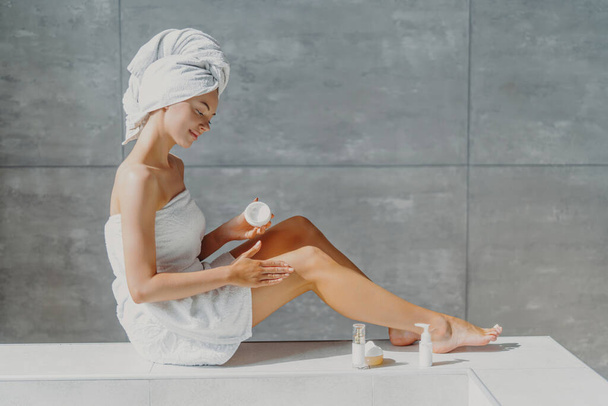 Pretty woman applies moisturizing cream, slender legs. Beauty routine in bathroom. Softness after lotion. - Photo, Image