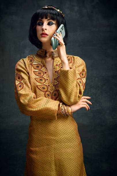 Portrait of young woman with expressive makeup in image of Cleopatra talking on mobile phone against dark vintage background. Concept of antique culture, history, comparison of eras, art, beauty, ad - Foto, Imagen