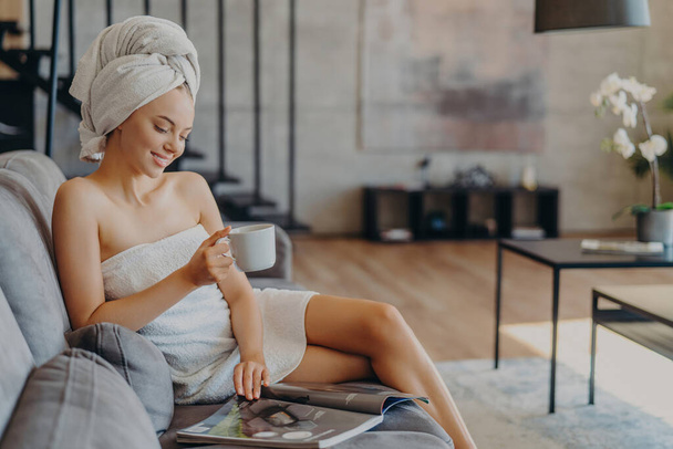Entspannte Frau mit gesunder Haut genießt die Zeit zu Hause. Zeitschriften lesen, Kaffee schlürfen, in Handtücher gewickelt. Erfrischende Wellness-Freizeit. - Foto, Bild