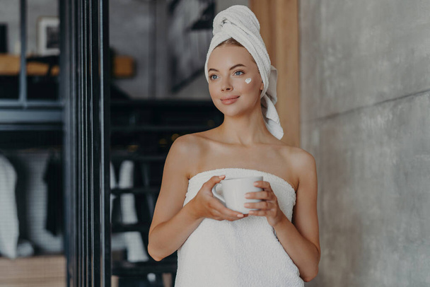 Thoughtful model with cream on face, towel wrapped, holds tea, undergoes beauty treatments indoor - Photo, Image
