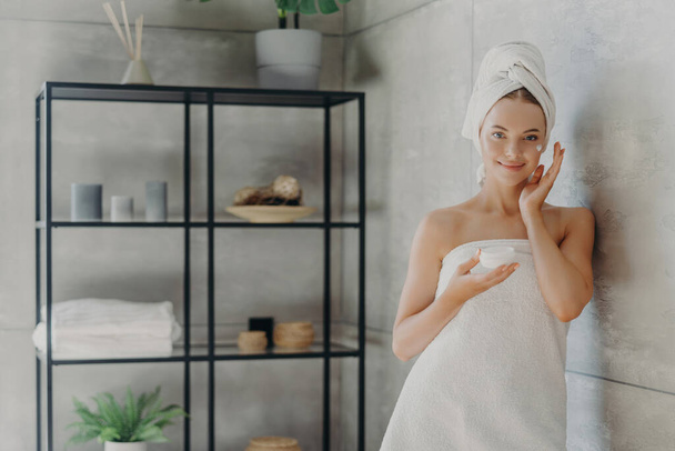 Modelo saudável usa creme, contém frasco cosmético, envolto em toalha, perto da parede do banheiro acolhedor. Cuidados da pele - Foto, Imagem
