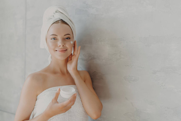 Lovely model applies moisturizer, holds cream jar, beauty routine, towel wrapped, poses against grey wall - Photo, image