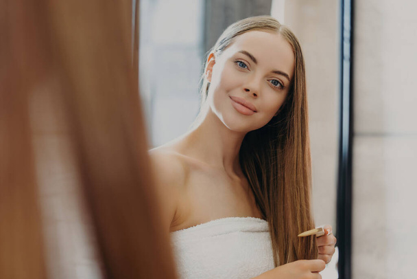 Pretty woman with minimal makeup, full lips, combs hair in mirror, wrapped in towel, self-care for fabulous look. Hair care concept - Foto, imagen