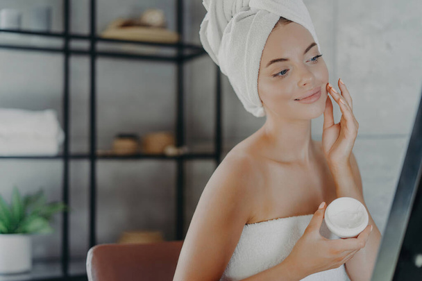 Pleased woman applies face cream, admires reflection, towel-wrapped, anti-aging after shower for healthy skin. Skin care concept. - Photo, Image