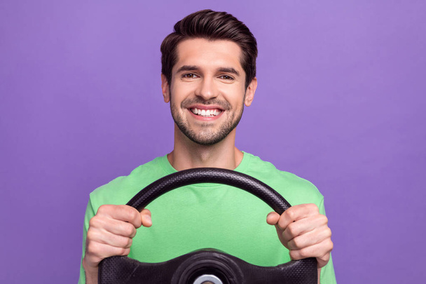 Portrait of satisfied cheerful guy with bristle wear green t-shirt holding steering wheel driving car isolated on violet color background. - Фото, изображение