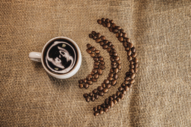 Świeże palone ziarna kawy w postaci W-iFi i gorąca filiżanka kawy w worku jutowym na stole .  - Zdjęcie, obraz