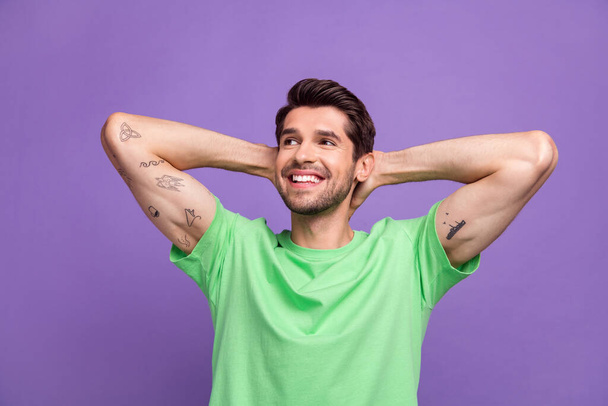 Portrait of good mood positive guy with bristle wear green t-shirt arms behind head look empty space isolated on violet color background. - Zdjęcie, obraz