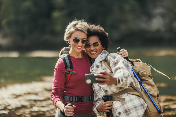 Two mixed-race female friends taking selfies by a river during a hike along the mountain. - Photo, Image