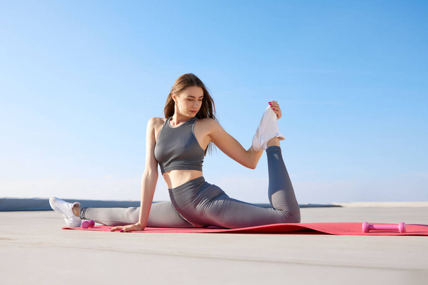 Flxible, sportive young woman in comfortable sportswear training on fitness mat outdoors on warm summer day. Stretching. Concept of sport, active and healthy lifestyle, beauty, fitness, ad - 写真・画像