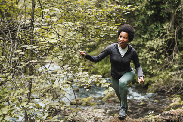 成熟したアフリカ系アメリカ人の女性ハイカーは、山の中を探索しながら川を渡り、屋外トレーニングを楽しむ. - 写真・画像
