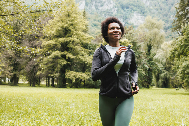 Beautiful smiling mature African American woman is running through green grass near the mountain landscape. - 写真・画像
