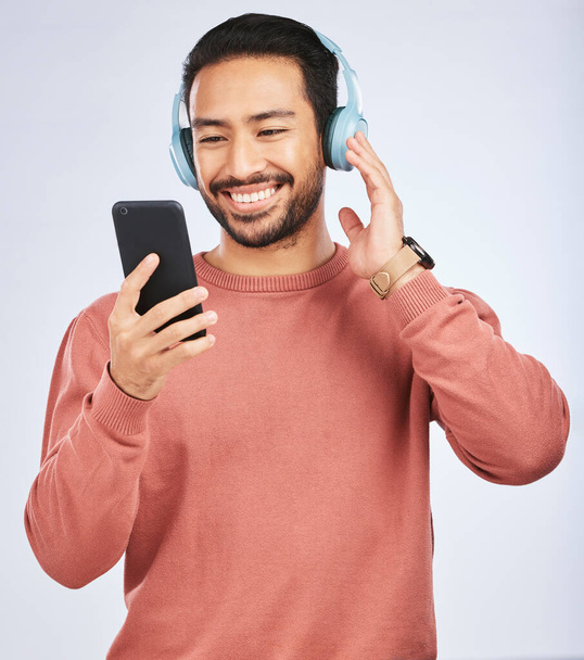 Man, headphones and smartphone for music in studio, white background and podcast app. Happy asian male model, cellphone and listening to audio, streaming mobile sound and online radio media with tech. - Foto, imagen
