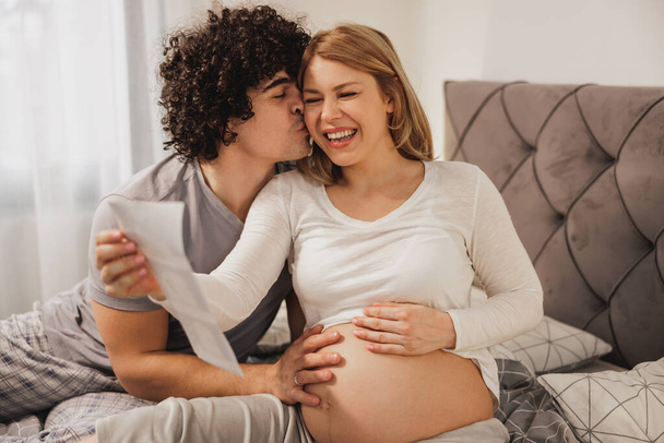 Feliz pareja embarazada mirando el escaneo médico de su bebé mientras se relaja en una cama en el dormitorio. - Foto, Imagen