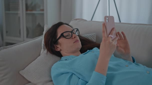 赤い髪の女性は、スマートフォンのテキストメッセージを使用してソファに横たわっているか、ソーシャルメディアでニュースフィードをスクロールします - 映像、動画