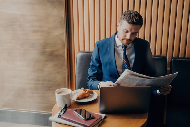 Gericht ondernemer in formele kleding leest financiële kroniek, koffiepauze met cappuccino, croissant, laptop open. - Foto, afbeelding