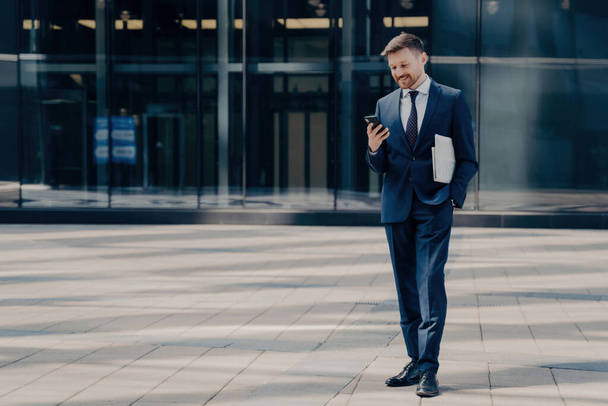 Cheerful employee in blue suit texts/chat on smartphone, holds newspaper, stands next to glass building outdoors. Business people and technology concept. - Photo, Image
