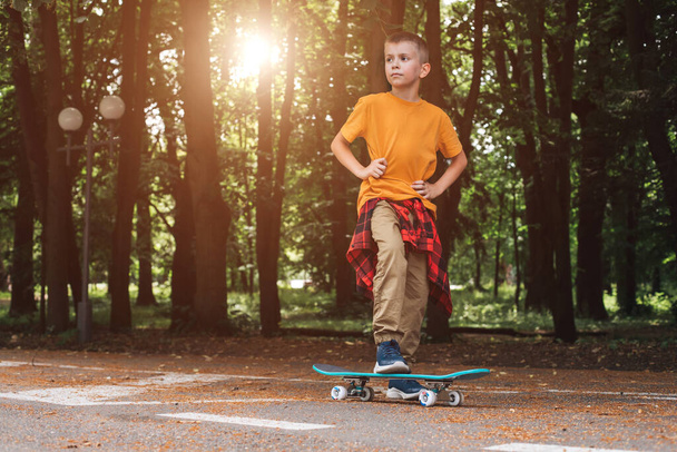 Szabadtéri tevékenységek gyerekeknek. Egy vidám fiú egy gördeszkán marad a játszótéren. Fehér iskolás fiú sétál. Előnézet - Fotó, kép