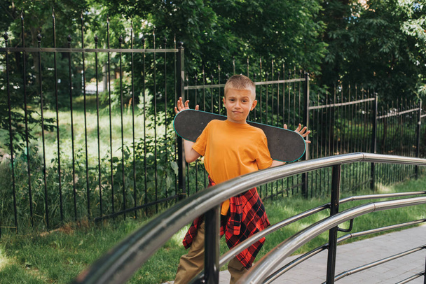 Aktivitäten im Freien für Kinder. Ein fröhlicher kleiner Junge in heller Kleidung bleibt mit einem Skateboard stehen. Kaukasischer Schuljunge bei einem Spaziergang. Frontansicht - Foto, Bild