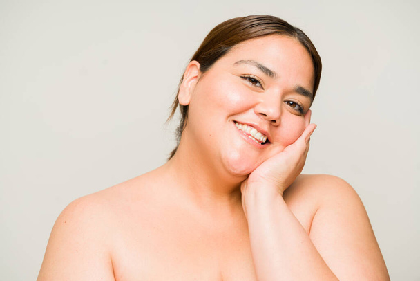 Attraktive übergewichtige Frau lächelt und nimmt Blickkontakt auf, während sie ihre weiche Haut berührt, nachdem sie ihre Hautpflege-Routine erledigt hat - Foto, Bild