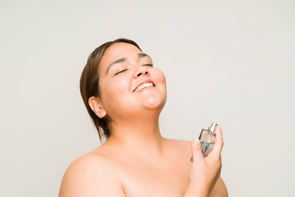 Aantrekkelijke ontspannen vrouw die glimlacht terwijl ze parfum spuit en er gelukkig uitziet met veel eigenliefde en zelfvertrouwen - Foto, afbeelding