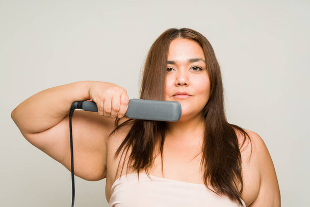Plus size hispanic woman doing a new hairstyle and using a straightening iron while looking at the camera isolated in a studio background - Photo, Image