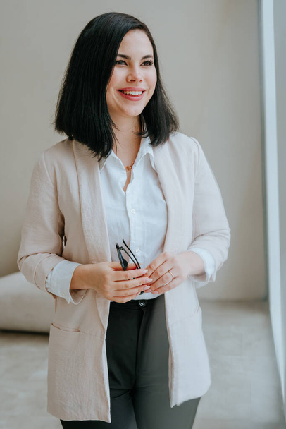 Portrait of young smiling brunette businesswoman holding glasses against a light wall indoor. Selective focus. - Foto, Imagem