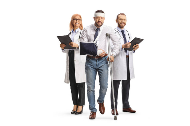 Team of doctors and a man with an injured head and arm standing with crutches isolated on white background - Fotoğraf, Görsel