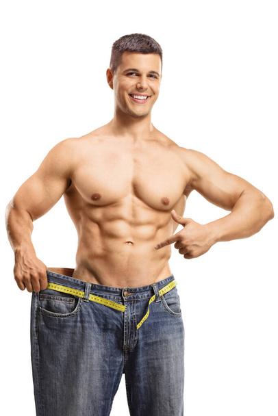 Fit young man trying on a big pair of jeans and pointing at his waist isolated on white background - Fotoğraf, Görsel