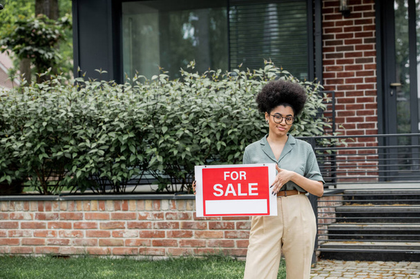 glücklich afrikanisch-amerikanischer Immobilienmakler hält Schild mit zum Verkauf stehenden Schriftzügen in der Nähe von Haus in der Stadt - Foto, Bild