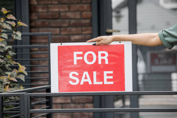 oříznutý pohled na africký americký realitní makléř držící cedulku s nápisem na prodej u plotu domu - Fotografie, Obrázek