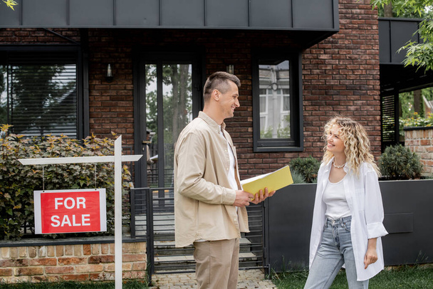 cheerful property realtor with folder talking to woman near modern cottage in city - Photo, Image