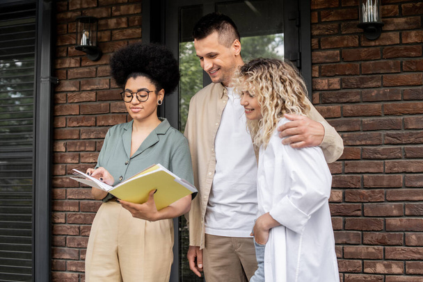 agente inmobiliario afroamericano mostrando documentos a los compradores encantadores abrazos cerca de nueva casa - Foto, imagen