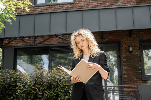 stylish blonde real estate agent looking at documents in folder near contemporary urban building - Photo, Image