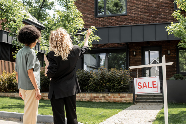 vista posteriore di bionda agente immobiliare che punta con mano, mostrando cottage in vendita alla donna afro-americana - Foto, immagini
