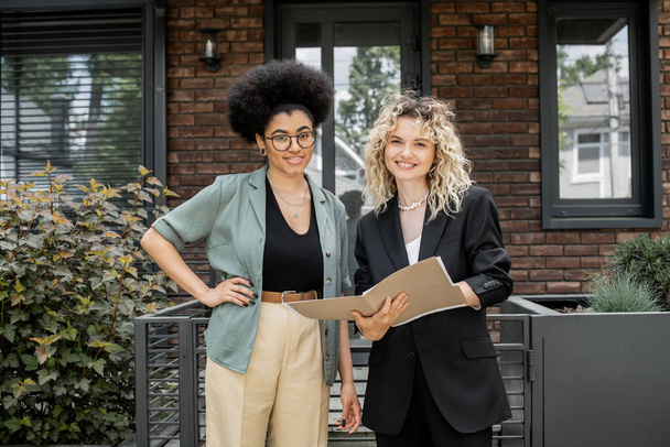 happy realtor with folder and african american woman with hand on hip looking at camera near house - Fotografie, Obrázek