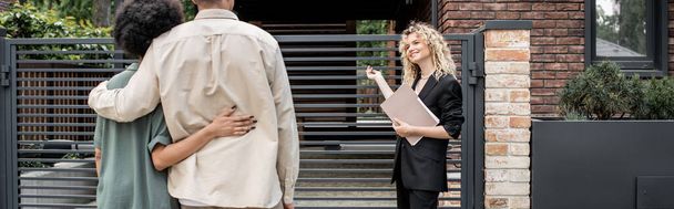 agent immobilier blonde positive avec dossier pointant vers une nouvelle maison près d'un couple multiculturel, bannière - Photo, image
