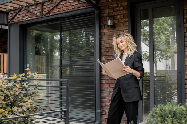 smiling blonde real estate agent looking at documents in folder near contemporary new house - Foto, Imagem