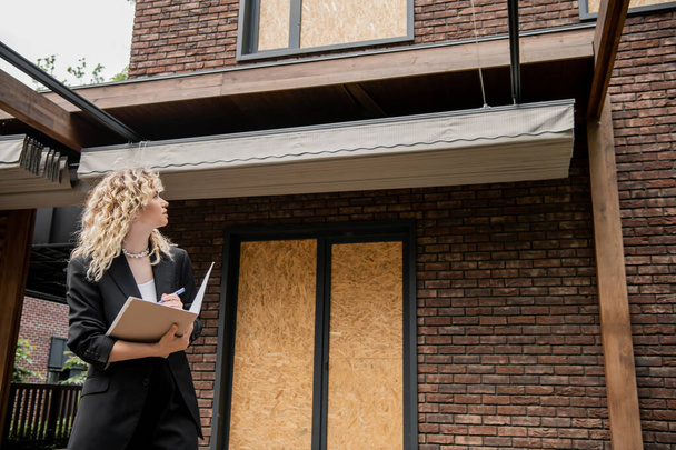 блондинка агент по недвижимости глядя на новое здание и писать в папке на открытом воздухе - Фото, изображение