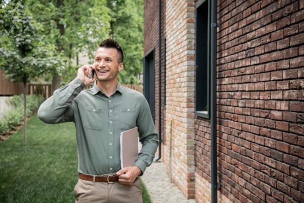 pleased real estate agent with folder looking away while talking on mobile phone near cottage - Photo, Image