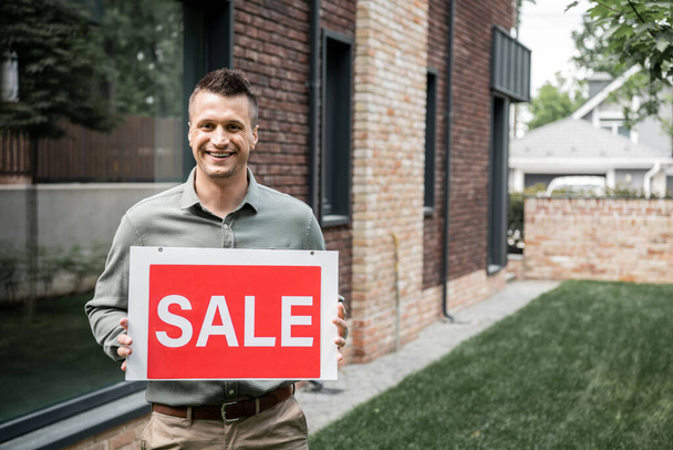 optimistic realtor holding signboard with sale lettering and looking at camera near cottage - Photo, Image