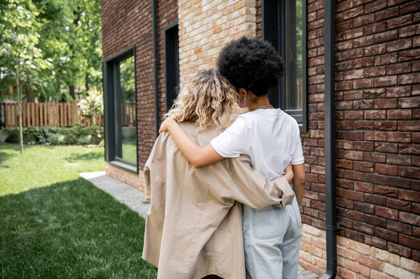 back view of multiethnic lesbian couple embracing near new private cottage outdoors, happy owners - Photo, Image