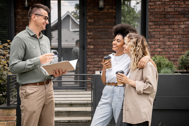 cheerful multiethnic lesbian couple with coffee to go talking with real estate agent near new house - Photo, Image