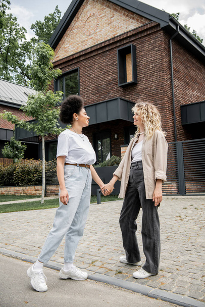 longitud completa de la pareja lesbiana multicultural tomados de la mano y sonriendo el uno al otro cerca de su propia casa de campo - Foto, Imagen