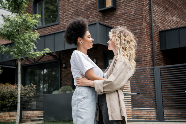 side view of interracial lesbian couple embracing and smiling at each other near new own cottage - Photo, Image