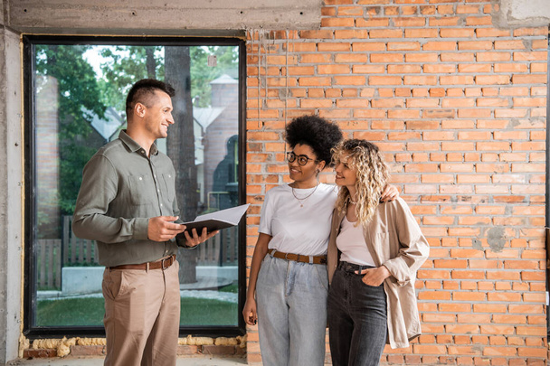 smiling real estate agent with folder talking to multiethnic lesbian couple inside new cottage - 写真・画像