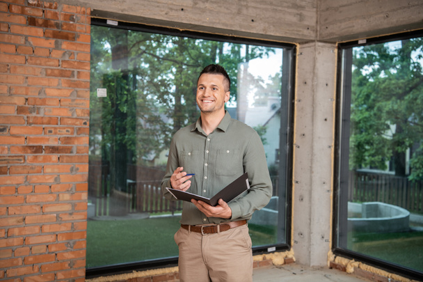 happy successful real estate broker holding folder and pen while standing inside new cottage - Photo, Image