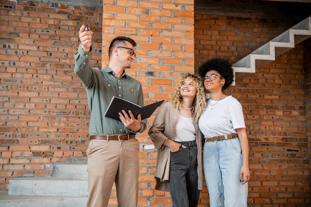 real estate agent pointing with hand near smiling interracial lesbian women in new cottage - Photo, Image